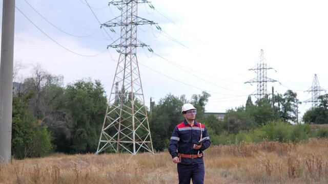 Electrical Power Worker Engineer In Helmet Working With Digital Tablet, Near Tower With Electricity