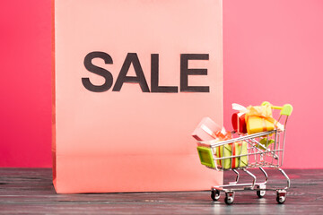 sale lettering on paper bag near small trolley with gifts on pink, black friday concept