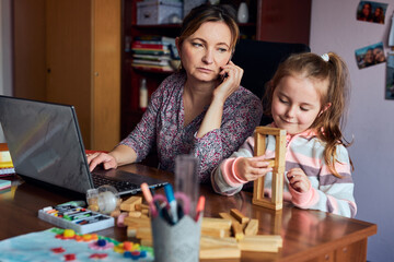Fototapeta premium Woman mother working doing her job remotely during video chat call stream online course webinar on laptop from home while her daughter playing with bricks toy
