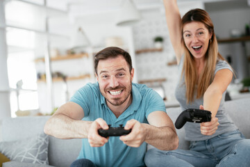 Naklejka premium Husband and wife playing video game with joysticks in living room. Loving couple are playing video games at home