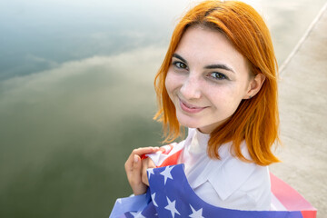 Happy smiling red haired girl with USA national flag on her shoulders. Positive young woman celebrating United States independence day.