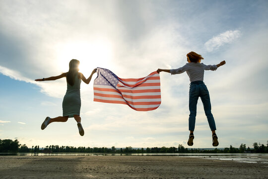 Two young friends women holding USA national flag jumping up together outdoors at sunset. Silhouette of girls celebrating United States independence day. International day of democracy concept.