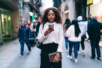 Beautiful dark skinned curly woman holding modern smartphone connected to 4G internet reading messages,african american female watching videos from social networks on city street holding mobile phone