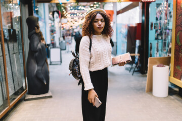 Half length portrait of african american female dressed in stylish outfit standing in store during free time, beautiful 20s dark skinned hipster girl looking at camera spending time in shopping mall