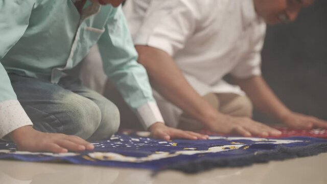 Muslim father and son praying or performing Salah while sitting on Prayer rug and touching head to mihrab or mosque printed on rug