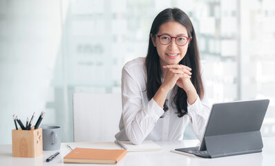 Portrait of young attractive asian woman with glasses relaxing while sitting at her office desk, smiling and looking at camera. - obrazy, fototapety, plakaty