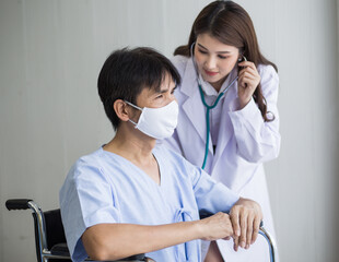 Asian woman doctor use a stethoscope to check the lung rhythm of a male patient who wear face mask while he sit on a wheelchair at hospital.