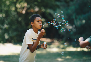 Cute african boy blowing bubbles outdoors with his friend and having fun on beautiful sunny day.