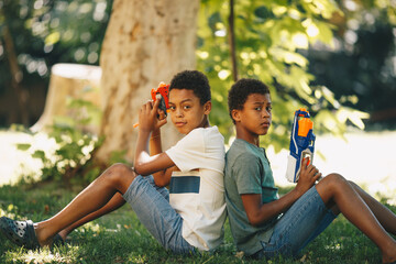 Two african brothers sitting outside on the lawn back to back and posing with water guns.