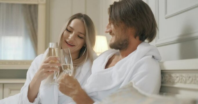 Affectionate couple toasting with champagne lying on their bed. Hotel, travel, relationships and happiness concept - smiling couple with champagne glasses in room
