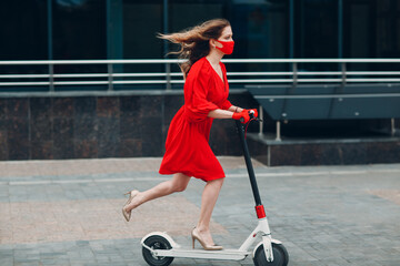 Young woman with electric scooter in red dress and gloves with face mask at the city. New normal fashion and coronavirus COVID protection concept.