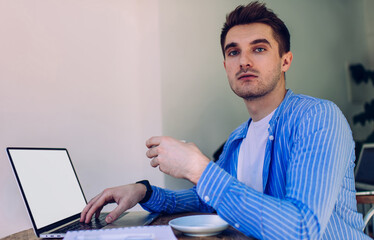 Fototapeta na wymiar Portrait of skilled male graphic designer with tea cup sitting a desktop table with mockup laptop and posing, Caucasian freelancer using blank netbook with copy space area for internet advertising