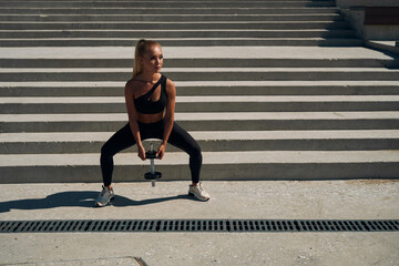 athletic blonde squats with dumbbells on the steps. young woman in black suit trains legs at sunrise