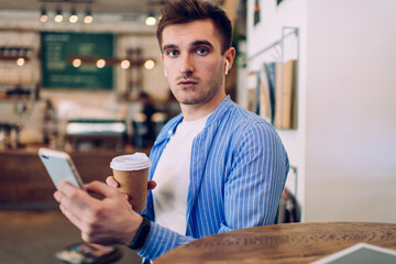 Portrait of good looking male millennial looking at camera while choosing audio songs in electronic headphones, handsome hipster guy with coffee to go listening music on radio station using mobile app