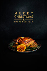 Baked whole chicken with oranges and rosemary. Tray with a festive Christmas dish on a black table and black background with text - Merry Christmas and Happy New Year. Low key. Vertical. Copy space