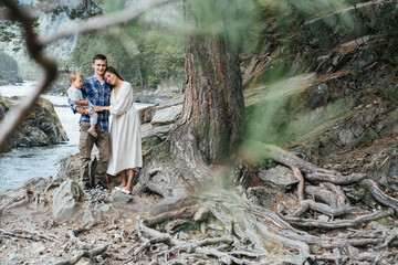 Young family father, mom and baby boy standing on the roots under big tree near mountain river