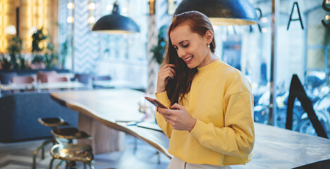 Happy hipster girl using cellphone device for mobility during online networking and browsing websites, cheerful woman in casual wear checking wifi connection in loft cafeteria for sending media file