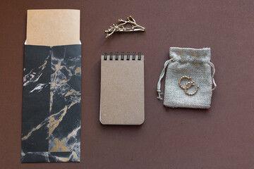 marble envelope brown notebook and golden earrings on small canvas bag and tree branch shaped decoration on brown background. Stationery mock up