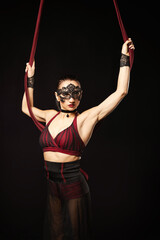Brunette beautiful caucasian woman in Lace mask doing circus tricks on red silks isolated on black background.