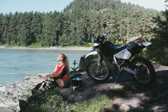 Female enduro biker wearing helmet and motoshoues sitting with her motorcycle on the cliff near mountains river