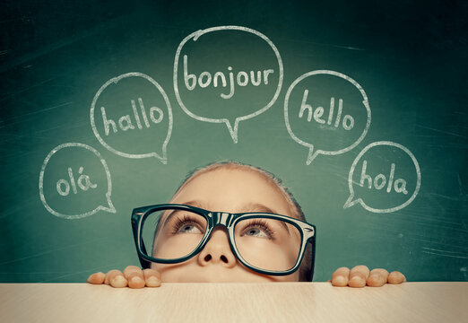 Beautiful cute little girl with eyeglasses hiding under table and looking at hello word in french, english, spanish, portuguese and german in speech balloons. K-12 foreign language learning concept.