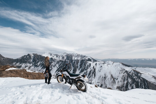 Woman motorcyclist standing near her enduro motorcycle on snowy mountain top, snow peaks skyline view, high beautiful mountains