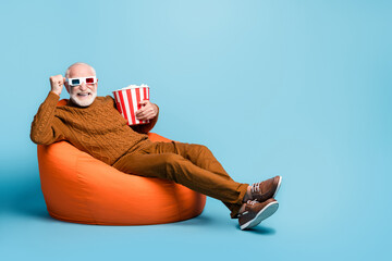 Portrait of his he nice attractive funky cheerful cheery glad bearded grey-haired man sitting in bag chair eating corn watching film having fun isolated over blue pastel color background - obrazy, fototapety, plakaty