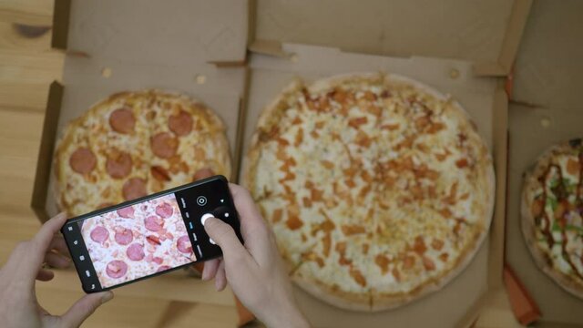 Female Food Blogger Taking Photos Of 3 Pizza By Smartphone In Pizzeria. Photographing Food For Social Network. Woman Hands Taking Picture of Delicious Pizza With Cellphone on Wooden Table in Kitchen.