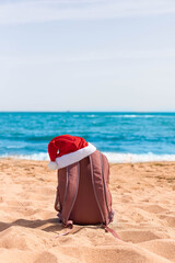 Santa hat on a sports backpack lying on the beach of the sea coast. Christmas beach holiday travel poster