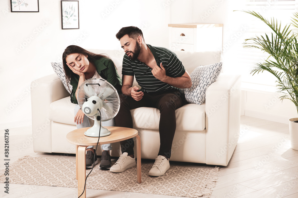 Wall mural couple suffering from heat in front of fan at home. summer season