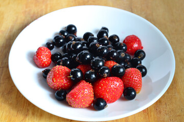 strawberries and black currants on a white plate and wooden table, harvesting, fresh and healthy vitamin-rich food