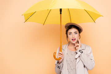 stylish woman in autumn outfit touching face under yellow umbrella on peach
