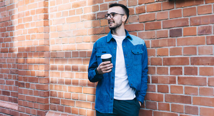 Positive caucasian male in casual denim wear and spectacles standing near brick wall on street, smiling 20s hipster guy recreating outdoors smiling and holding disposable coffee cup on leisure