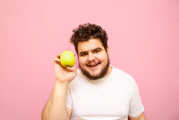Positive young overweight man isolated on pink background with green apple in hand, looking into...