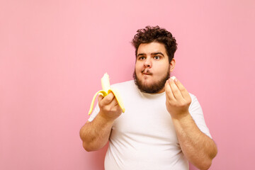 Funny curly fat man with a beard eats a banana on a pink background, looks away with a funny face....