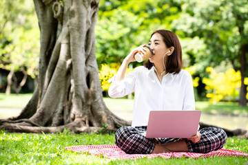 Asian woman holding cup of coffee with listening pop music from earphones and bright smiling face. Relax in the public park with laptop. Work from outside concept.