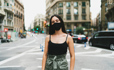 Beautiful stylish girl wearing medical face mask on a city street background. Young elegant hipster woman put on protective face mask outdoors. Urban fashion outfit during COVID-19 quarantine period.