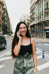 Cheerful caucasian hipster girl standing at the city street and talking on her smartphone. Young attractive woman in trendy outfit having phone conversation while standing on a city road background