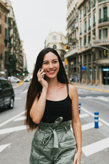 Fototapeta premium Cheerful caucasian hipster girl standing at the city street and talking on her smartphone. Young attractive woman in trendy outfit having phone conversation while standing on a city road background