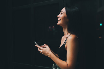 Side view photo of young caucasian female smiling aside while standing at the dark window with a smartphone in her hands. Positive hipster girl chatting with friends online by a mobile phone app.