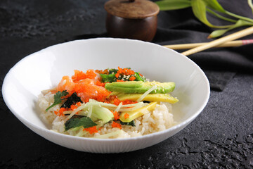Japanese cuisine. Rice with avocado, salmon, vegetables, caviar and herbs in a deep white plate on a black table. Restaurant menu. Background image, copy space