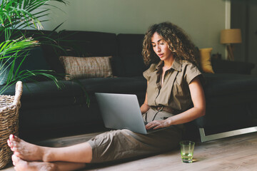 Beautiful female in casual outfit chilling on a floor beside comfortable couch in a living room with laptop on her knees. Web designer browsing  laptop while working on a remote project at home. - 382179575