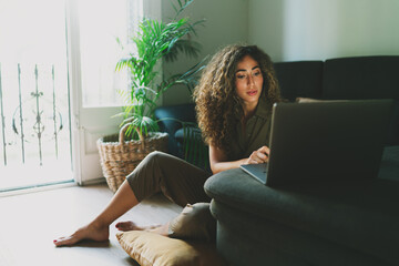 Beautiful hispanic woman working on a laptop computer while sitting on a floor at home during...