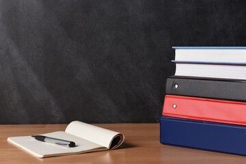 A stack of three different binders on desk in front of black board with two books and note pad and...