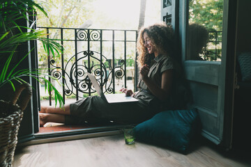 Side view photo of a cheerful young female in casual clothes sitting on a floor beside the open window and browsing laptop computer while spending time on a quarantine. Home office on self-isolation. - 382179964
