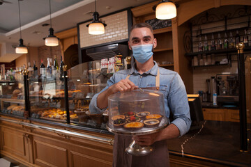Cheerful male baker wearing medical face mask, working at his coffee shop selling delicious...
