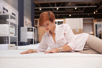 Gorgeous young woman examining orthopedic mattress, choosing new bed to buy