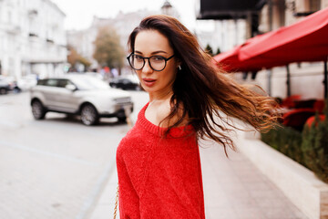 Fototapeta premium Lifestyle portrait of brunette woman with long hairs in red cardigan walking through street. Windy hairs. Stylish eyeglasses.