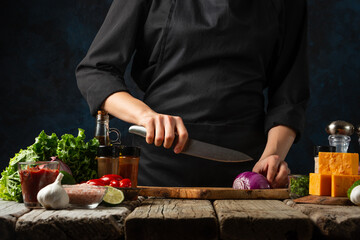 The chef in black professional uniform cuts with knife onion on the wooden board. Backstage of cooking tacos. Tasty mexican cuisine. Concept cooking recipe.