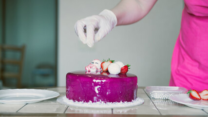 Obraz na płótnie Canvas A female hand in a glove decorates a mousse cake with pink icing with a figurine of a little deer made of white chocolate and berries.